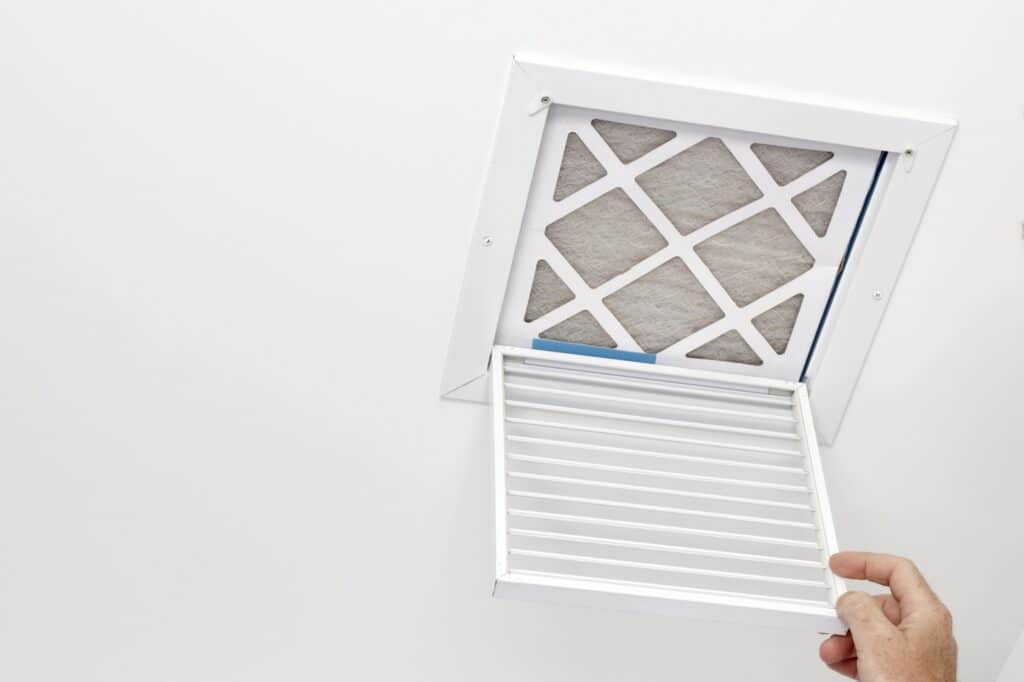 A small square HVAC system air intake vent reveals a dirty air filter in the ceiling. A man has opened a vent grill cover to reveal the dirty ceiling air filter.